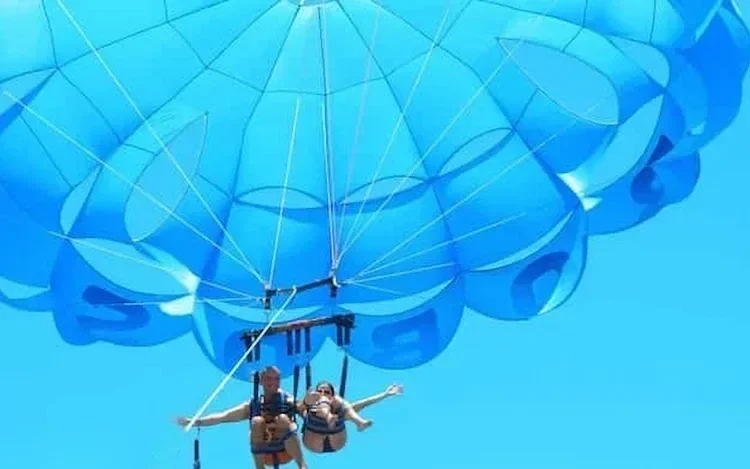 Looking straight up at a bright blue parasail canopy with two riders below, arms outstretched.