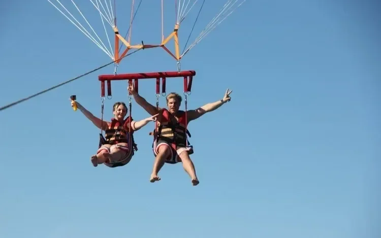 Two riders in life vests grin and wave from a parasailing harness against a clear blue sky.