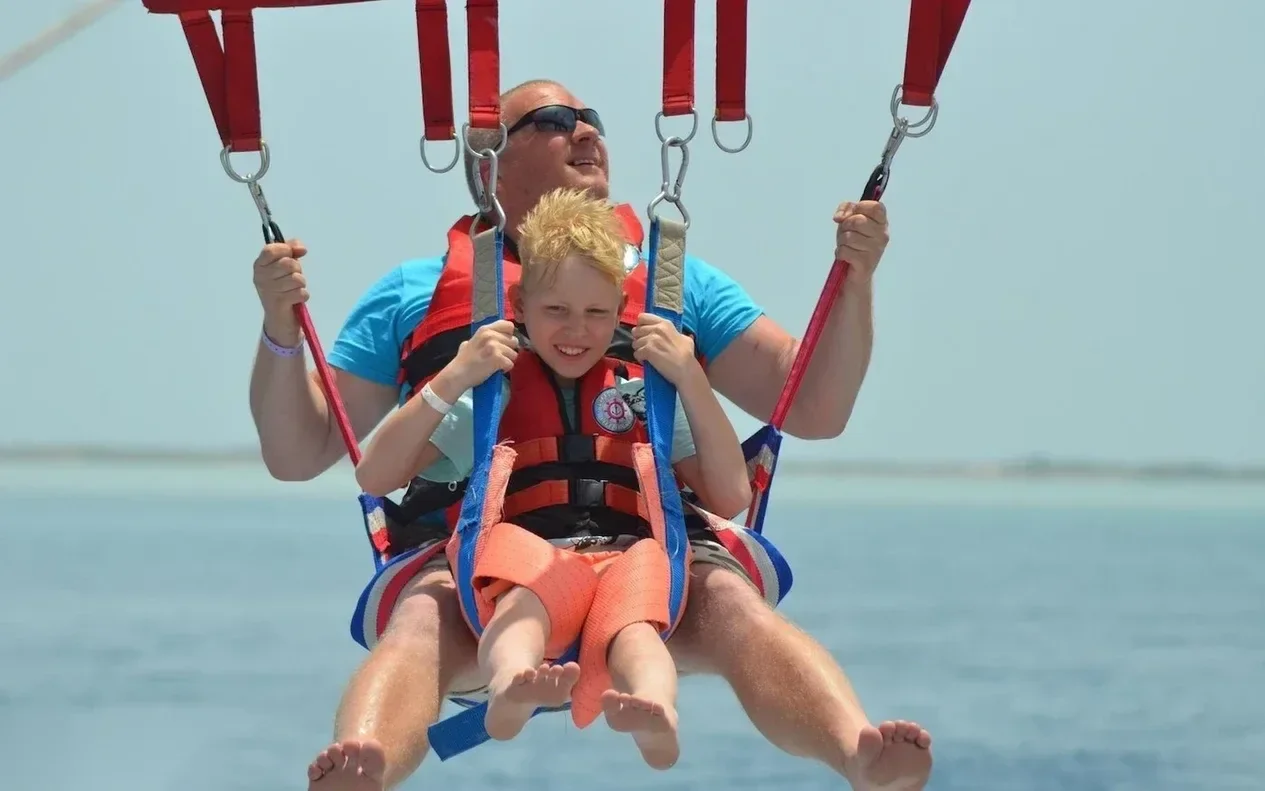 A father and young son ride tandem in a parasailing harness, both barefoot and smiling.