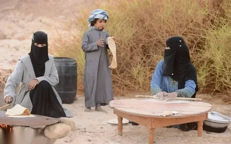Two Bedouin women in black niqabs and a boy in a headscarf making traditional flatbread over an open fire and on a low wooden table outdoors.