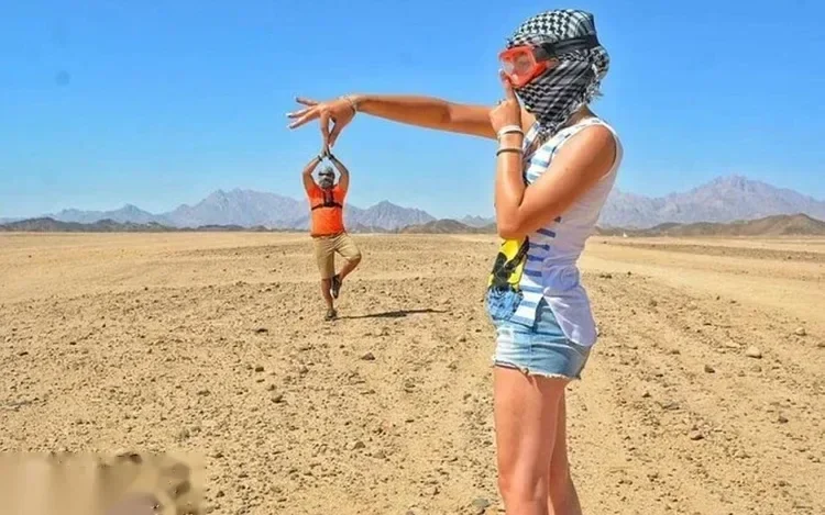 Woman wearing a snorkel mask on her head watches a man doing a jump pose in the flat desert with mountains in the background.