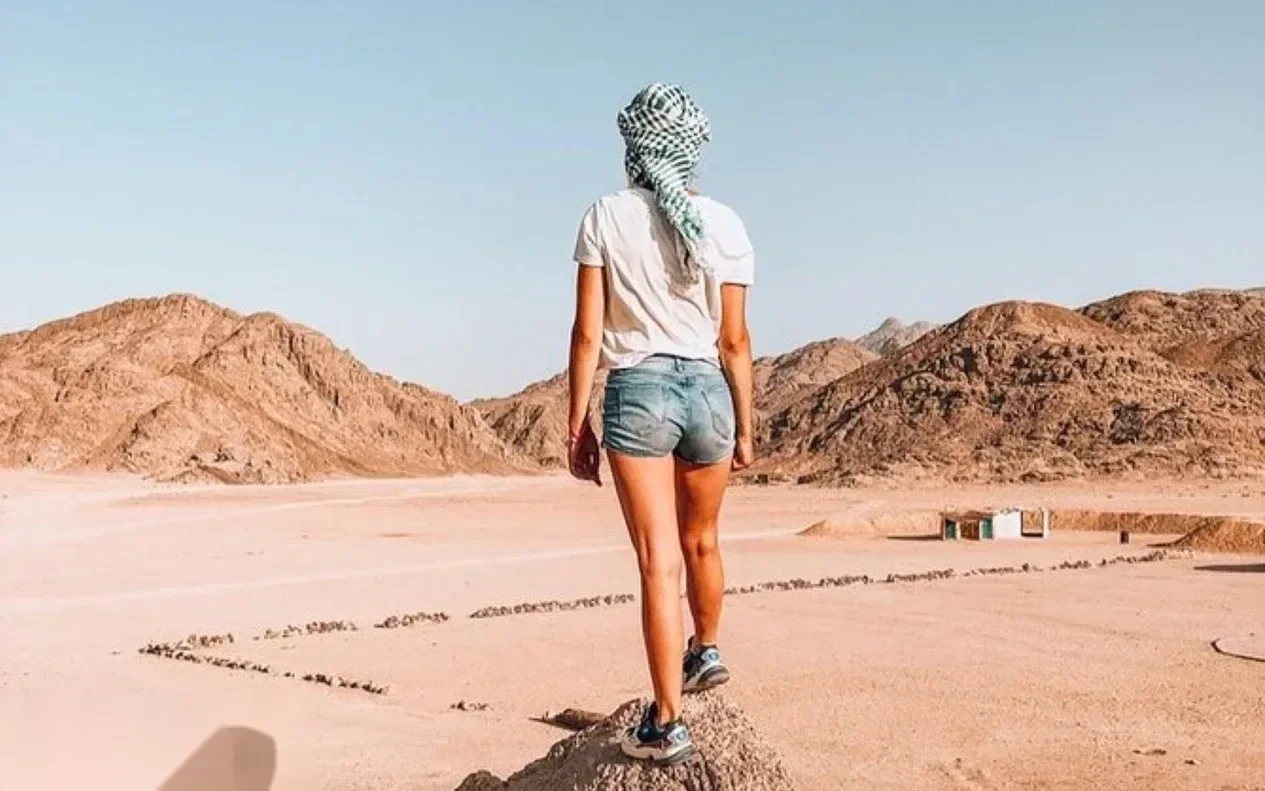 Woman wearing shorts and a headscarf walking away from the camera across flat desert ground with rocky mountains on either side.