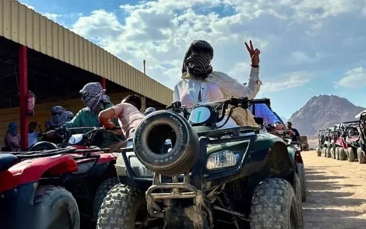 Rider wearing a checkered headscarf raises a peace sign while sitting on a quad bike among a line of other quad bikes at a desert camp.