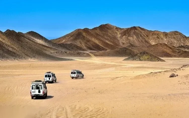 Three white 4x4 vehicles driving in convoy across an open desert valley surrounded by dark mountains.