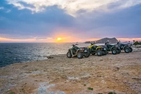 A row of quad bikes parked on a coastal bluff at sunset, with the sea and a small island visible beyond.