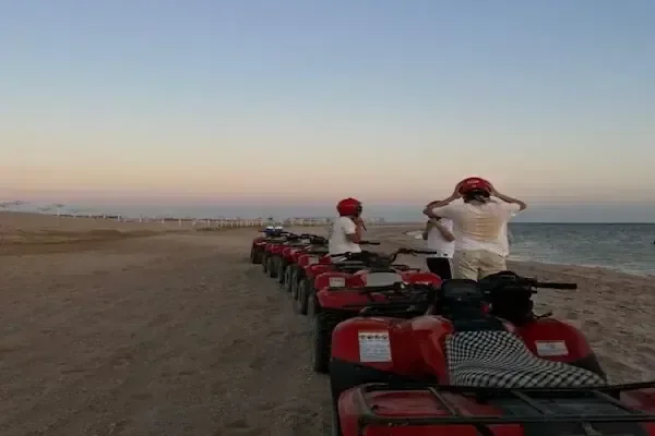 Two riders wearing red helmets stand beside red quad bikes on a sandy beach at dusk.
