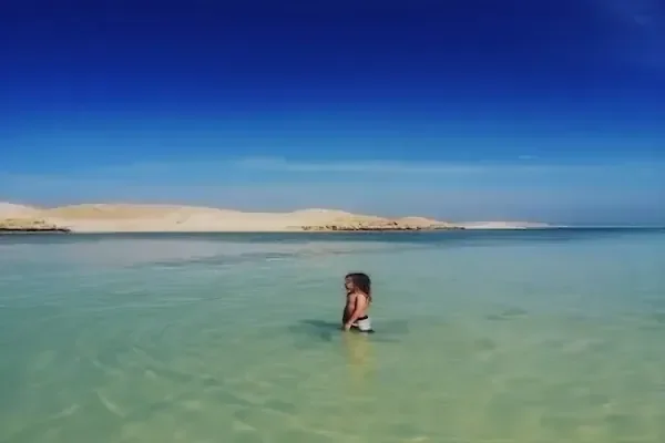 A woman wades into calm, shallow turquoise water with a large sand dune in the background.