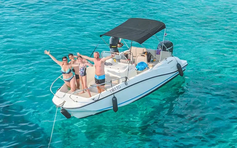 A group of four people stand on a moored speedboat with arms raised, smiling, in bright turquoise water.