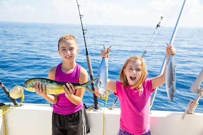 Two girls on a fishing boat proudly hold up their catches — a mahi-mahi and a smaller fish — with deep blue open sea around them.