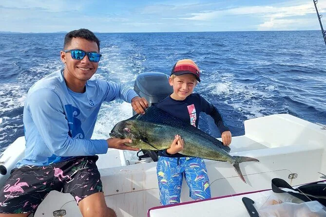 Two anglers stand at the stern of a boat, one reeling in a fish leaping from the water, with an ice-filled cooler of catch nearby.
