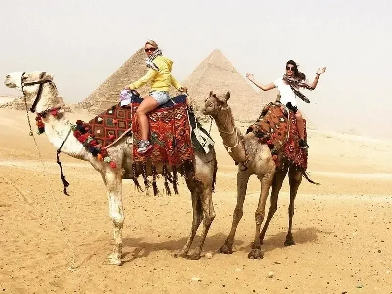 Two women riding camels side by side with a pyramid in the background, one waving to the camera.