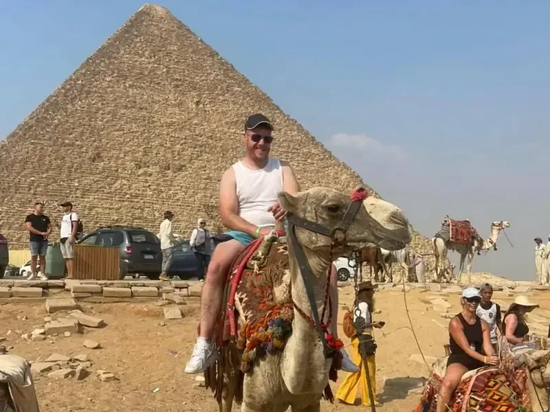 Man in a tank top riding a camel in front of a large pyramid, with other camels and parked cars nearby.