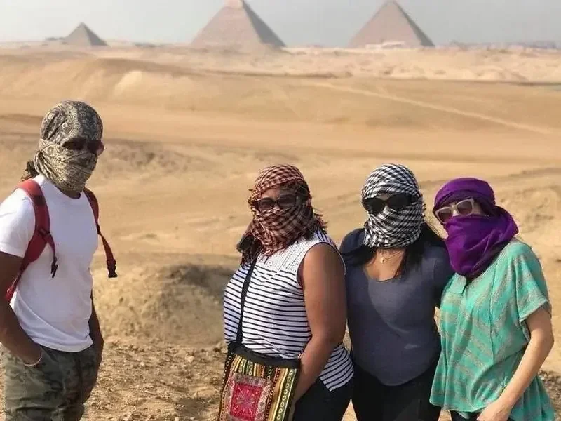 Four tourists with desert scarves wrapped over their faces standing with the Giza pyramids visible in the distance.