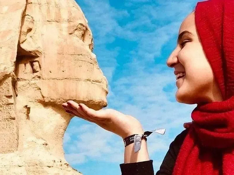 Close-up of a woman in a red headscarf smiling as she gently touches the chin of the Great Sphinx.