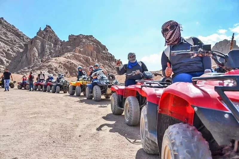 Line of red quad bikes with riders wearing desert scarves waiting in a rocky mountain valley.