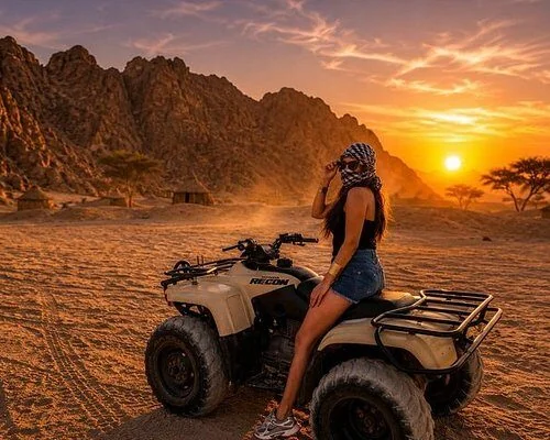 Woman in a shemagh scarf and shorts posing on a parked quad bike at sunset, with huts and acacia trees behind her.