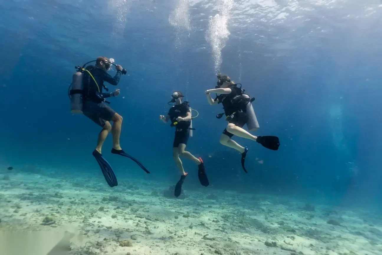 Three divers descend together mid-water above a sandy seabed, trailing bubbles toward the surface.