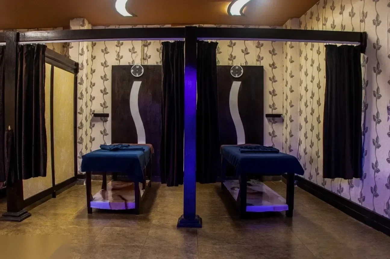 Dimly lit hammam treatment room with two massage tables facing each other beneath a purple LED accent light and curtained alcoves