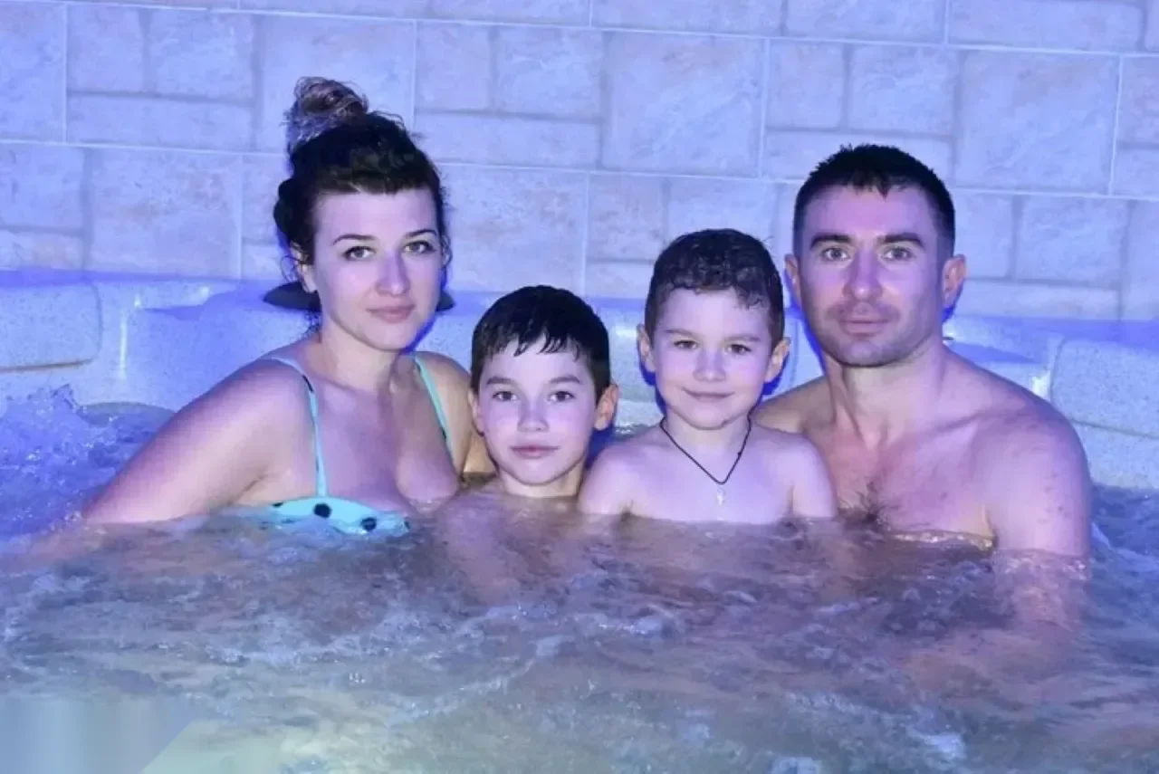 Family of four smiling together while soaking in a bubbling jacuzzi with tiled stone walls lit in purple light