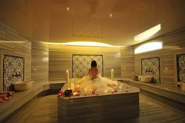 Woman wrapped in a white towel sitting alone in an ornate, candlelit marble hammam chamber decorated with rose petals