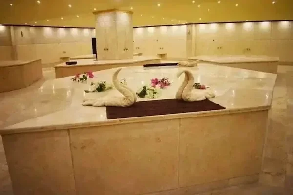 Woman wrapped in a white towel with a towel turban, reclining on a heated marble bench beside a carved stone basin under a classical relief carving