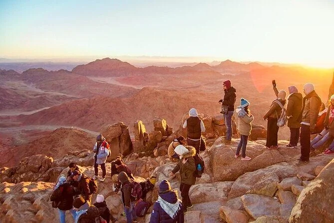 Group of bundled-up hikers standing on rocky outcrops watching the sun rise over layered desert mountains