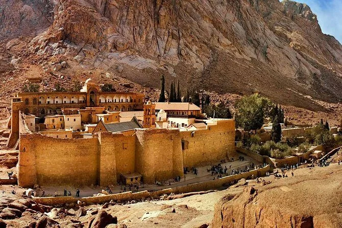 Wide view of Saint Catherine's Monastery with its fortified walls, domes, and cypress trees against a massive granite mountain