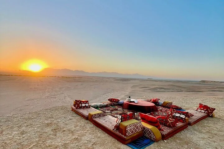 Bedouin-style desert majlis with red cushions and a round table set up on sand at sunset