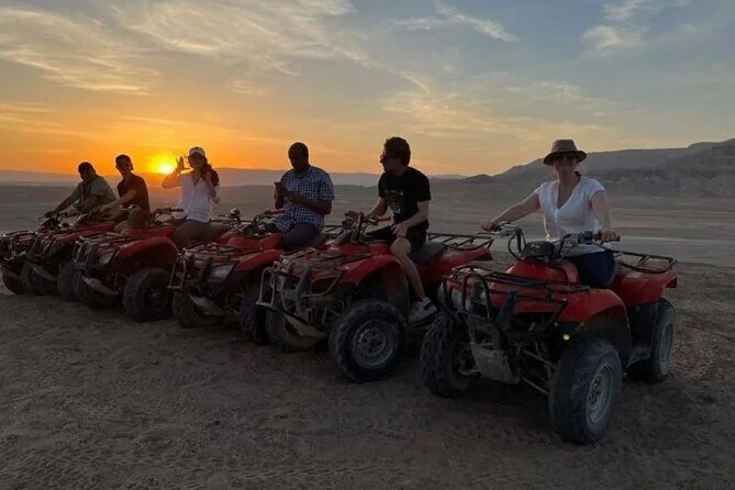 Six tourists sitting on red quad bikes posing together as the sun sets over the desert
