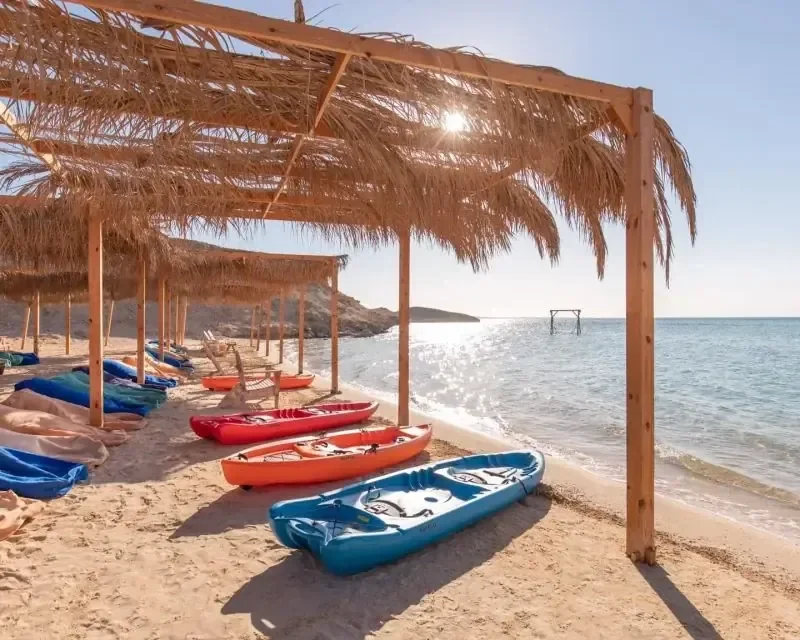 Colorful kayaks lined up on a sandy beach under straw-roof shade huts beside calm turquoise water