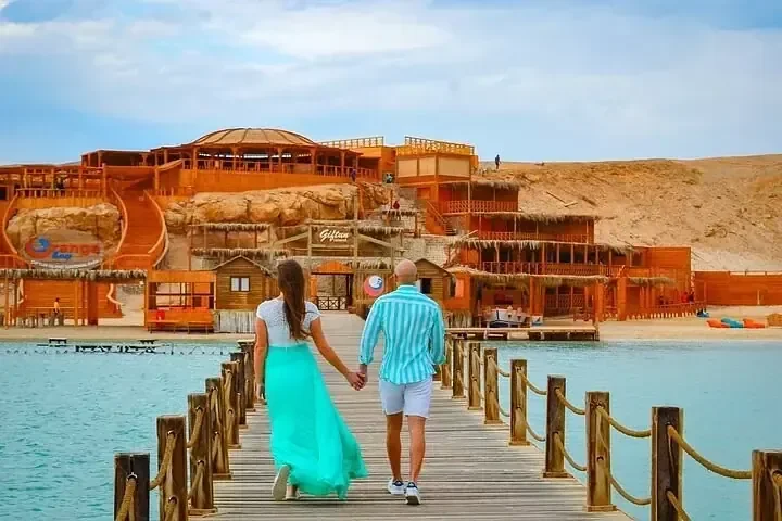 Couple walking hand in hand along a wooden pier toward a beach resort built over turquoise water