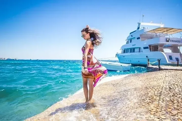 Woman in a flowing colorful dress walking along a stone jetty beside a docked yacht named Tiramisu