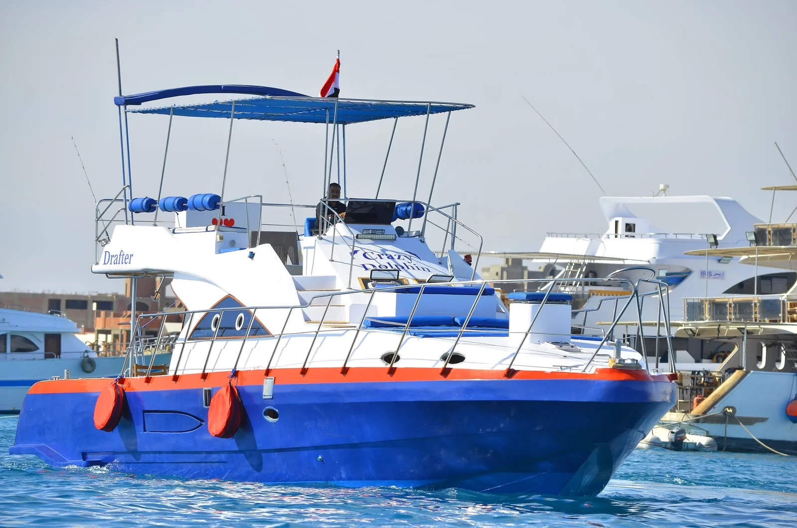Blue and white motor yacht named Drafter with Crazy Dolphin branding docked in a marina among other boats and waterfront buildings