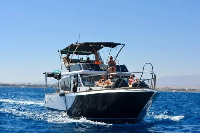 Black and white motor yacht cruising at sea with several tourists relaxing and sunbathing on the upper deck, coastline visible in the distance