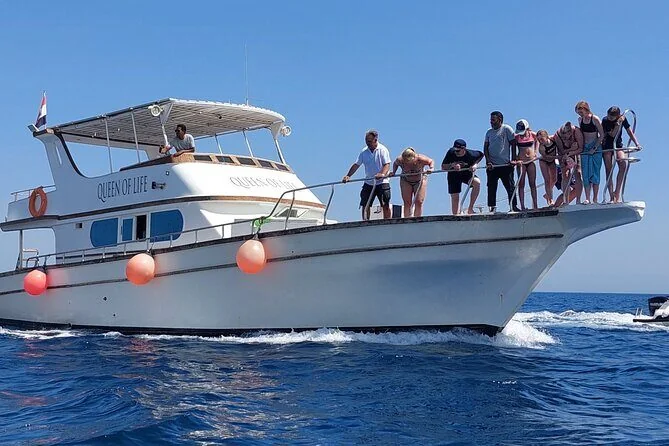 White yacht named Queen of Life with a group of tourists in swimwear gathered at the bow railing, orange buoys hanging from the hull